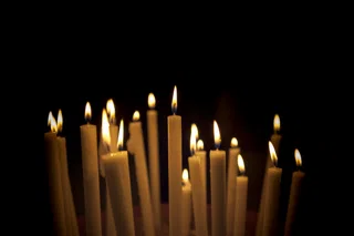 A row of beeswax tapers, lit, against a dark ground.